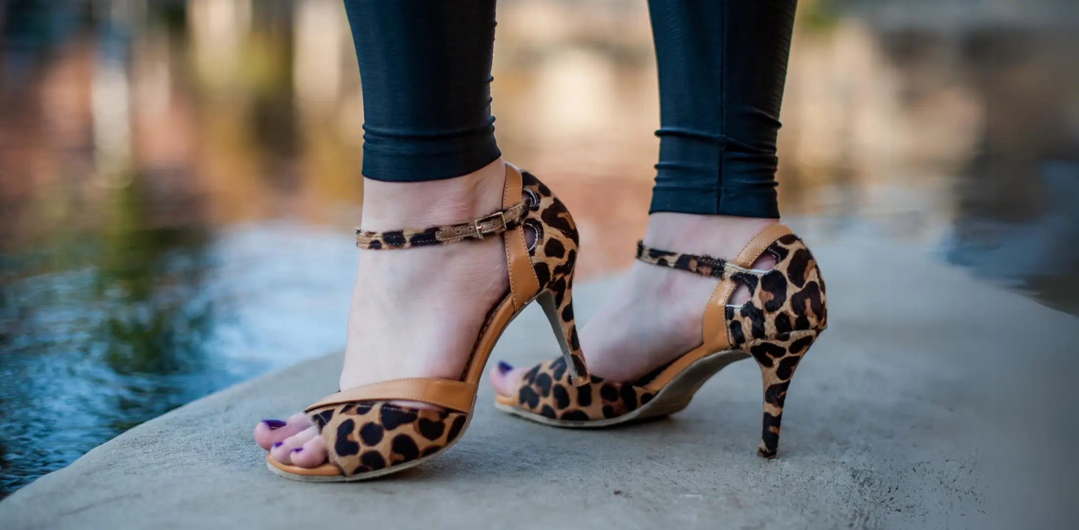 Woman standing on a ledge wearing leopard print high heel sandals with a water backdrop.