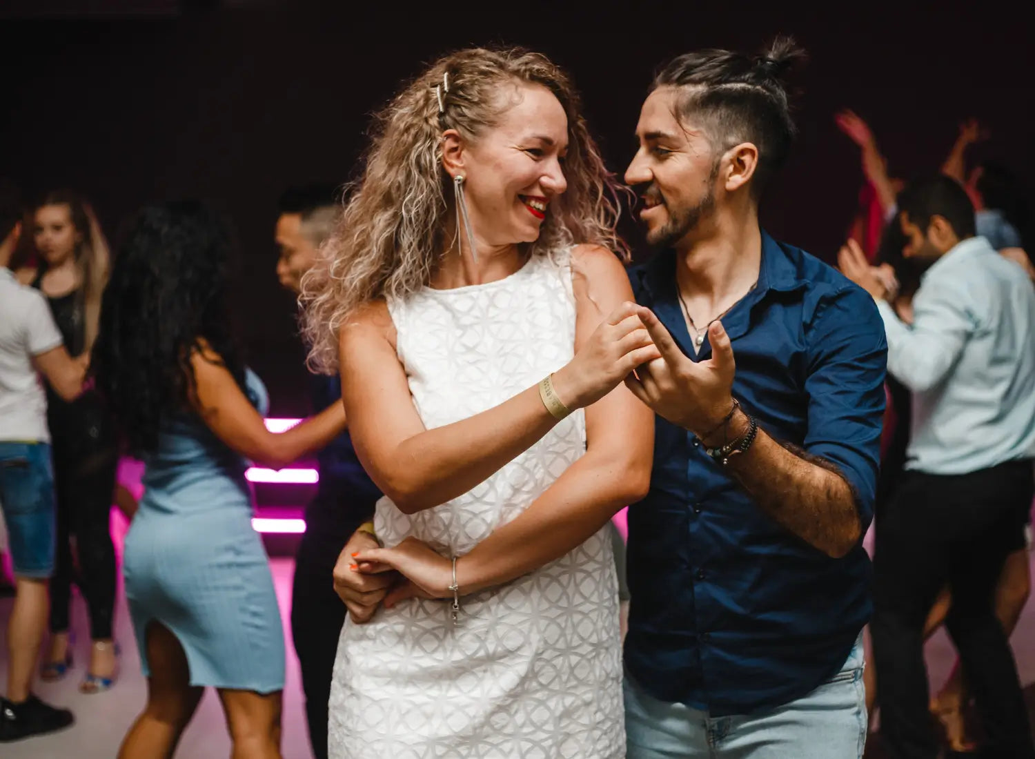 A man and woman smiling and dancing closely together on a lively dance floor with other dancers in the background.